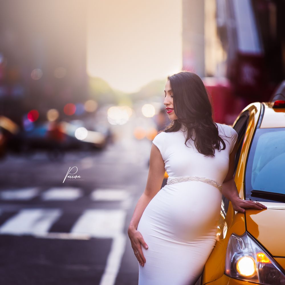 NYC Portrait of a pregnant brunette woman dressed in a tight white dress. She's on a New York City street next to an iconic yellow taxi cab.