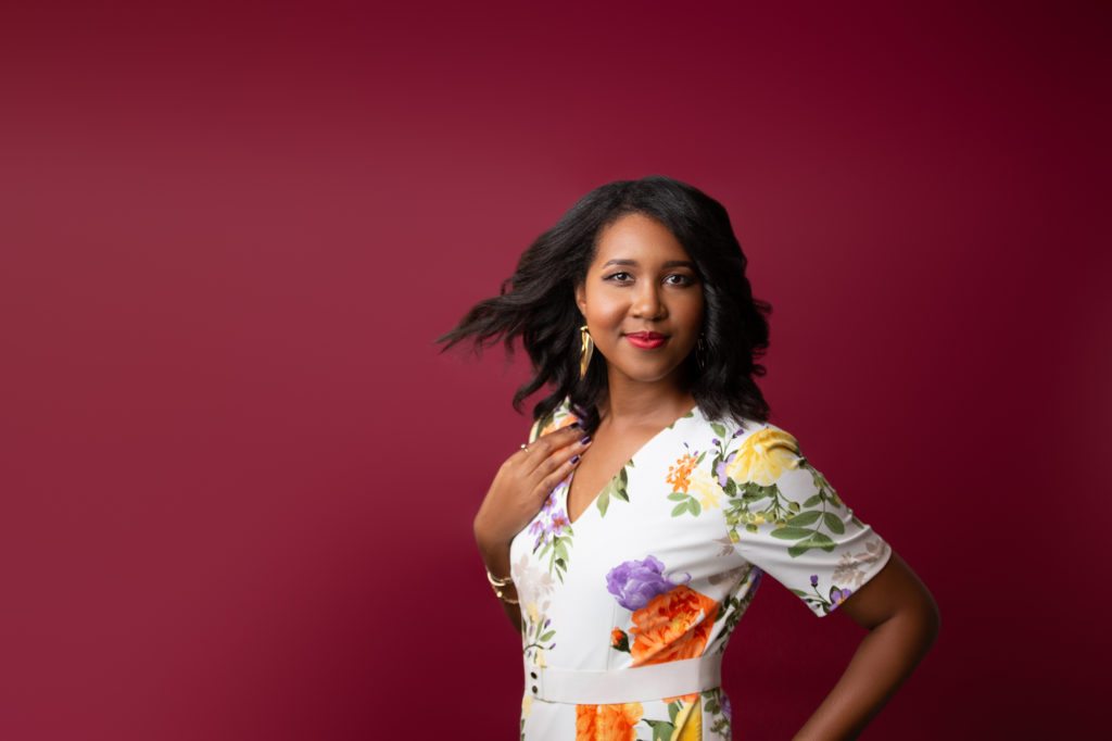 African- American woman with her hair loosely curled and swooped back. She has on a white floral shirt as she touches her left shoulder and is set on a bold red background.