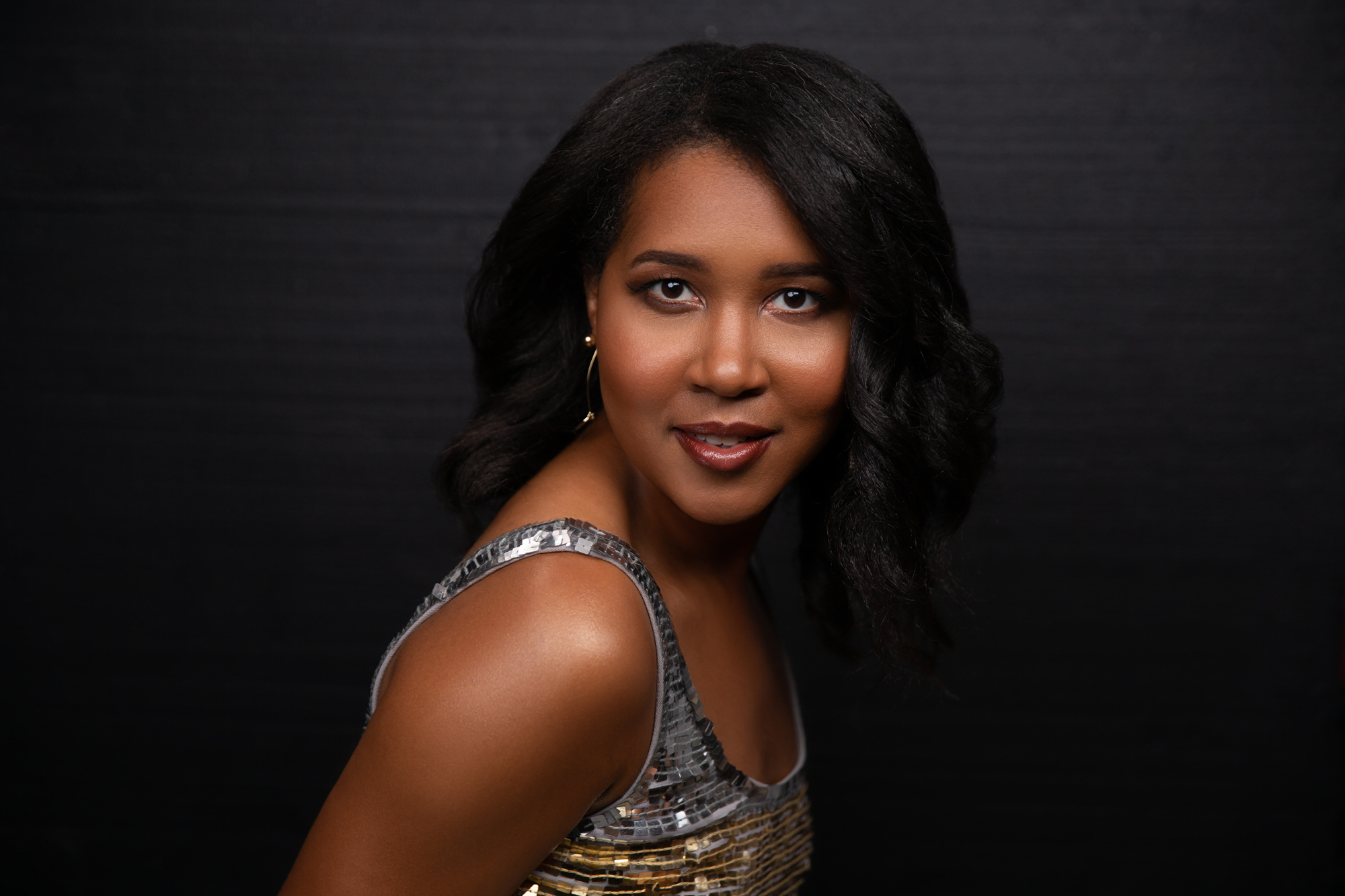 African -American Woman with her head tilted to the side - as she sits sideways in a gold/silver sequin dress. Her hair is adorning her shoulders and she has neutral make-up. She is smiling slightly and is on a black photo background