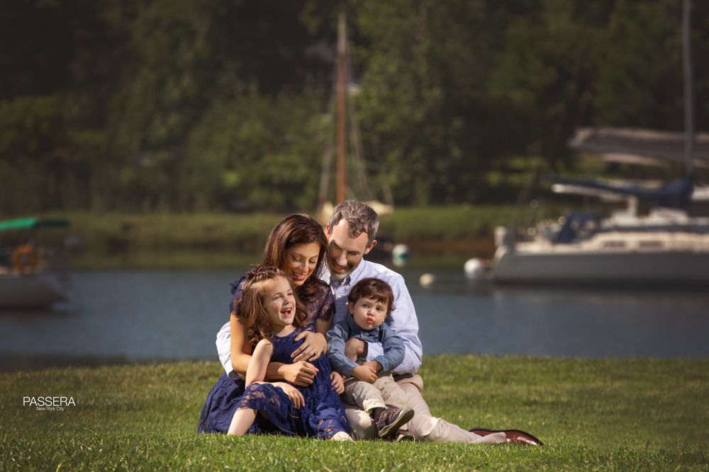 A family of a husband, wife, and 2 young children. They are sitting down on green grass near a lake in the back ground. The mother and father are embracing each other tightly why the both hold their young children and look at them lovingly.