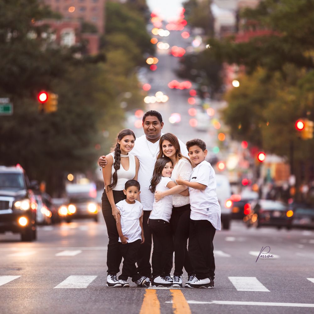 Family of Spanish decent all dressed in white t-shirts and black jeans with white sneakers. There is a mom, dad, 2 daughters and 2 sons. The y are standing in a city street cross walk with traffic blurred out in the background.